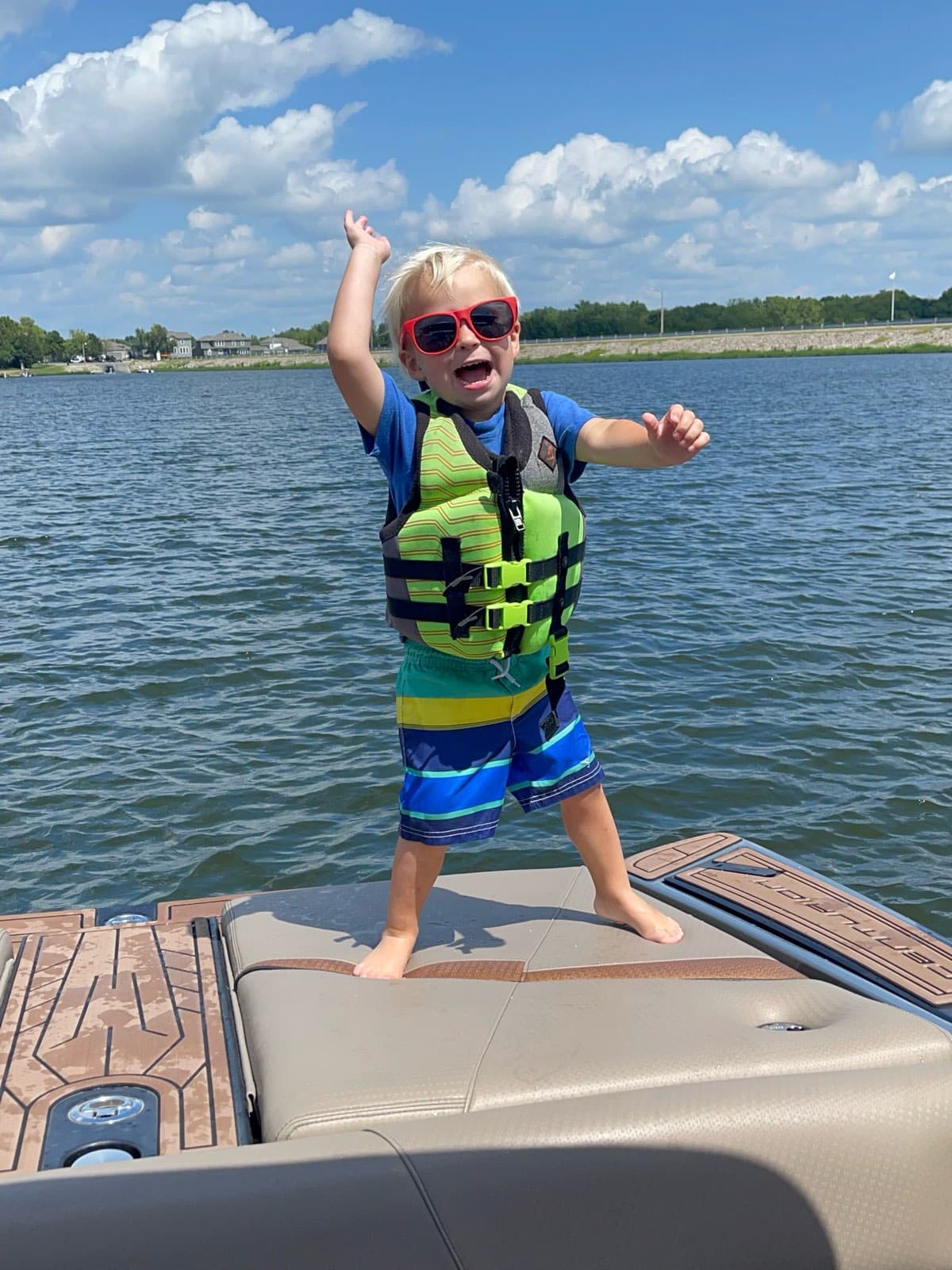 A toddler in a life jacket laughing on the bow of a pontoon boat, open lake behind